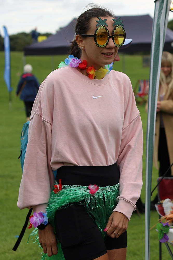 Woman wearing a pink Nike sweatshirt, large novelty pineapple sunglasses, a colorful flower lei, and a green grass skirt with flower decorations, dressed in a tropical-themed outfit at One More Lap for Kids' Village 2024.