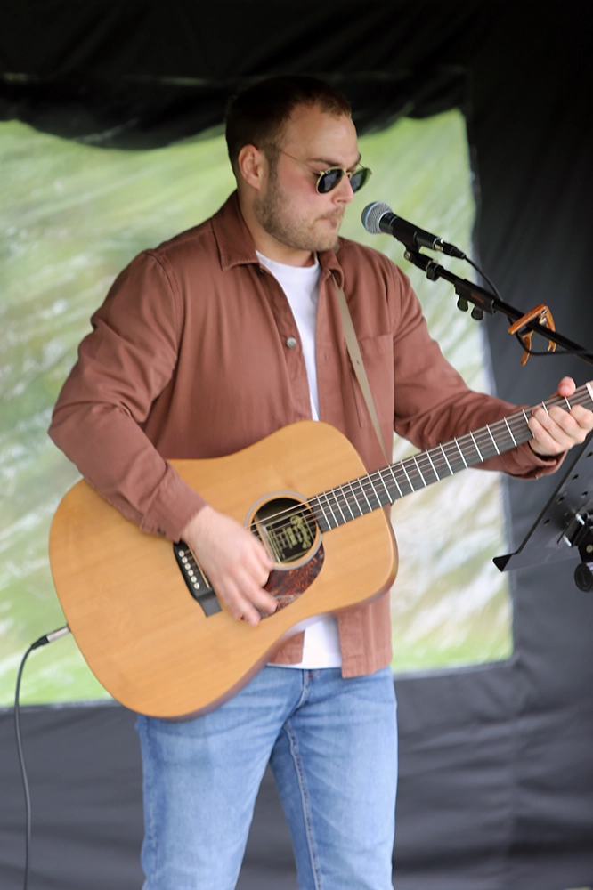 Male musician wearing sunglasses and a brown shirt, playing an acoustic guitar and singing into a microphone on an outdoor stage at One More Lap for Kids' Village 2024.