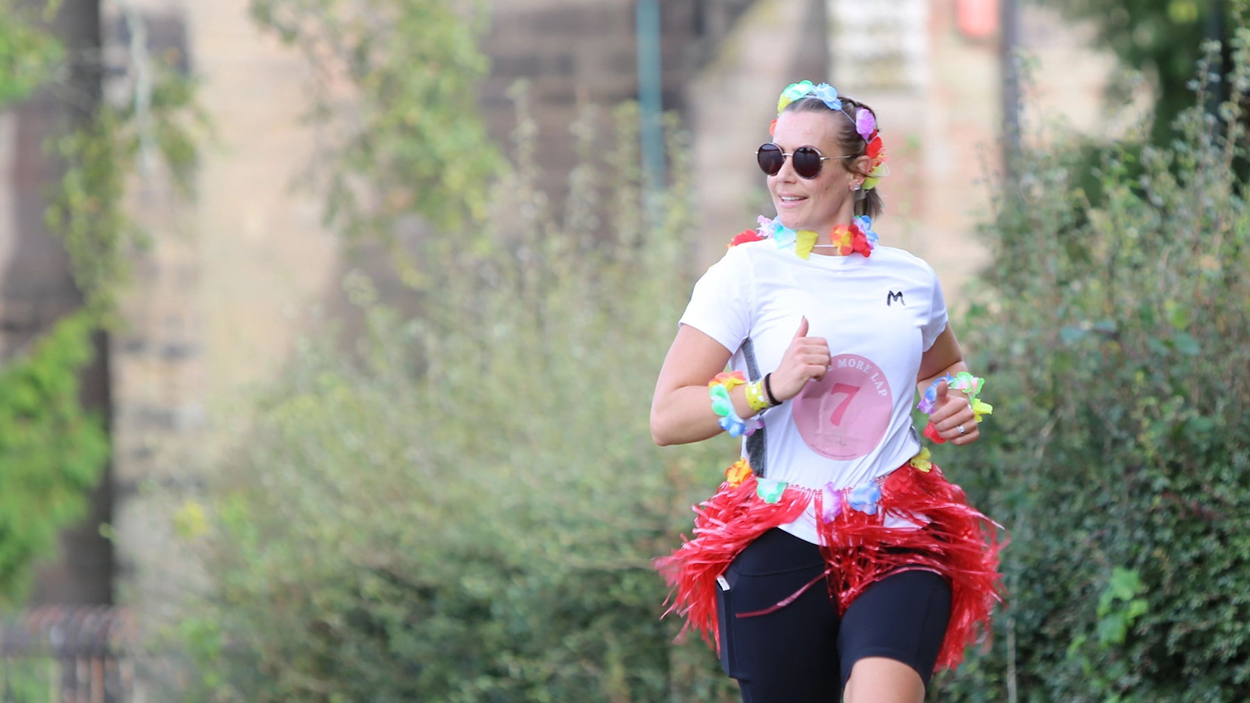 A smiling runner wearing sunglasses and a colorful flower lei, dressed in a white t-shirt with a red race number '7' and a red tinsel skirt. The runner has colorful flower bracelets and is running outdoors along a leafy path, with a stone wall in the background.