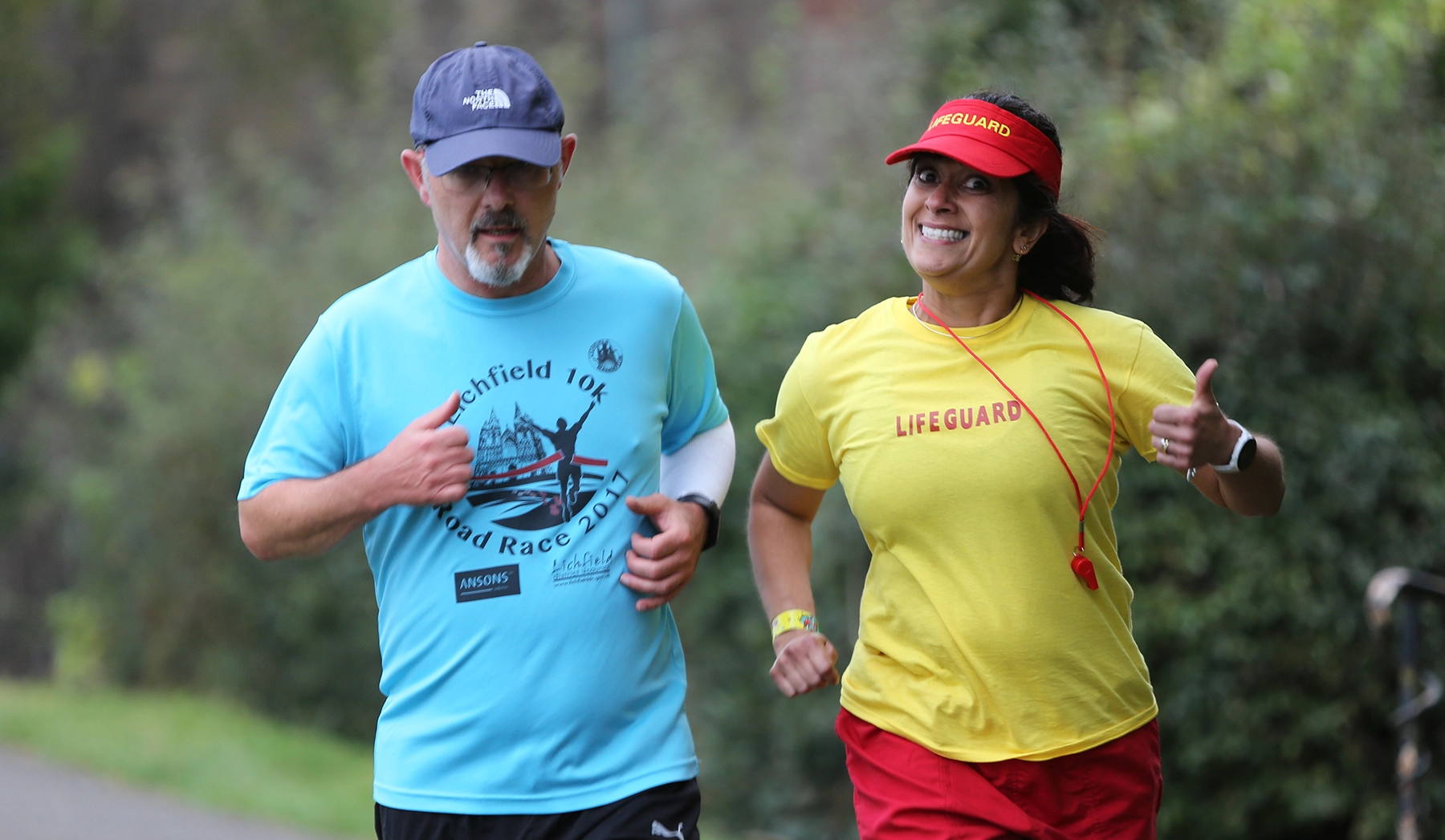 Man in a blue “Lichfield 10k Road Race” t-shirt and cap running alongside a smiling woman in a yellow “Lifeguard” shirt and red visor, both giving a thumbs up while jogging on a leafy outdoor path.