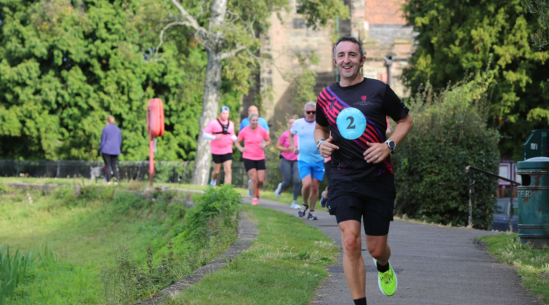 Man wearing a black and red running outfit with a race bib numbered 2 smiles while jogging on a park path, followed by a group of runners in brightly colored shirts, surrounded by lush greenery.