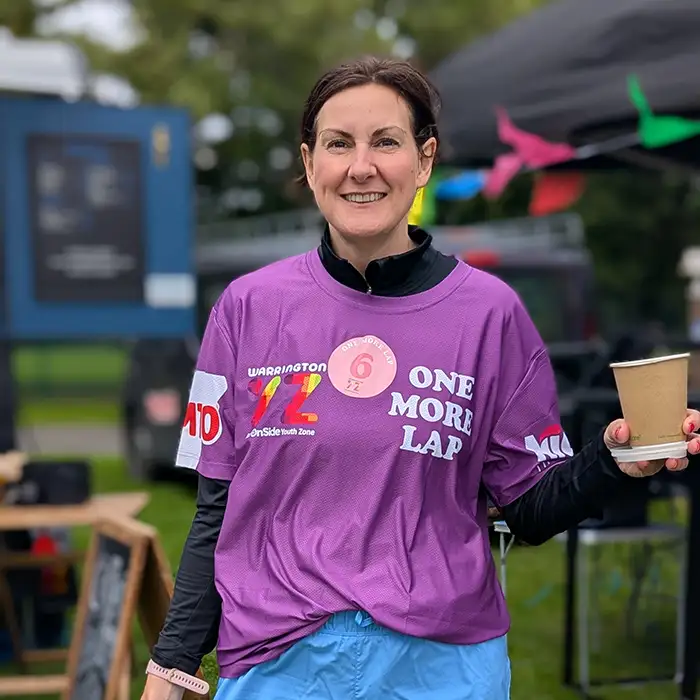 A smiling runner wearing a purple One More Lap T-shirt holds a paper cup, with colourful bunting and event tents behind her.