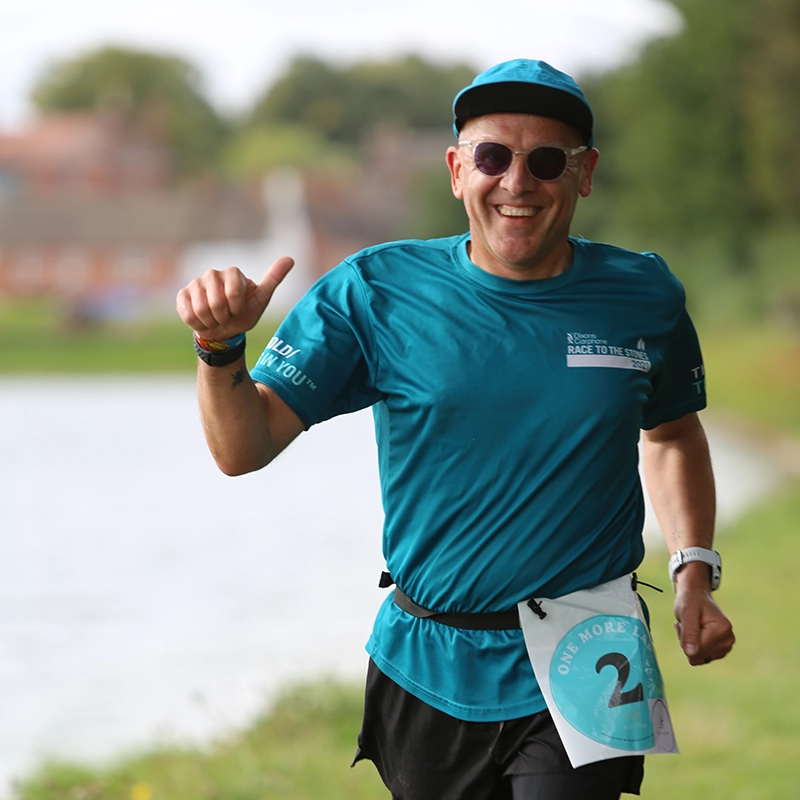 A smiling runner wearing a teal race shirt, matching teal cap, and sunglasses, giving a thumbs-up gesture while running along a riverside path. The runner's race number '2' is visible on their belt. The background shows a calm river, grassy bank, and blurred houses in the distance.